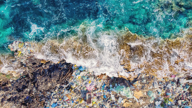 An aerial photo of a wave crashing onto a shoreline piled with marine debris.