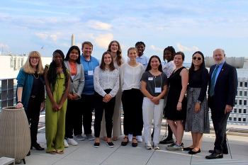 A group of 13 people, high school students and adults, smiling at the camera on a rooftop, with the Washington Monument in the background.