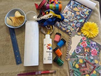 The supplies for the project laid out, including scrap ribbons, pom poms, a white crisp can, paint, and a paintbrush.