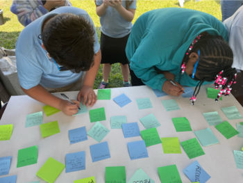 Two students lean over a table and write on sticky notes. 