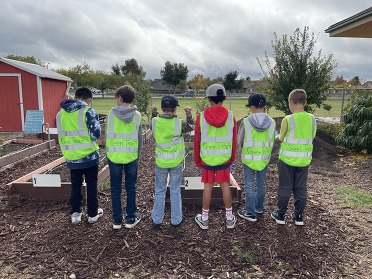 A line of students standing in a community garden while wearing yellow safety vests.