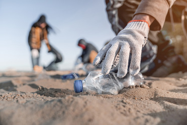 A hand wearing a glove that is picking up a plastic water bottle during a beach cleanup.