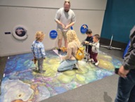 An adult overseeing children examining a floor mat that is a large photograph of a coral reef