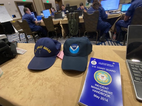 A NOAA hat next to a U.S. Coast Guard hat on a table in a room with responders on their computers.