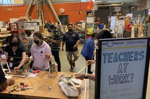 People in masks in a large room, sign reads TEACHERS AT WORK!