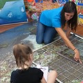 An adult pointing to a coral on a large mat picturing a coral reef, as a child nearby writes something on a clipboard