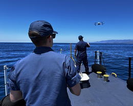 Two Coast Guard pilots stand at the end of a vessel with one launching an uncrewed aircraft by hand.