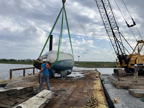 A blue boat is held upright on top of a dock by a crane while two workers in hard hats stand by. 