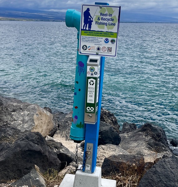 A long blue plastic recycling bin sits upon rocks next to the ocean. 