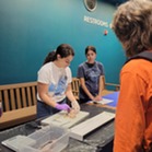 Woman looks down and holds a dissected fish, while two others look down at the fish. 