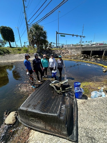 A group of people stand in shallow water in a concrete stream basin near large debris removed from a litter-trapping net. 