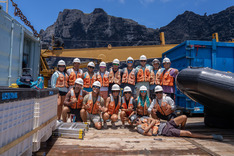 A group of people stand on the deck of a ship wearing life jackets and hard hats.