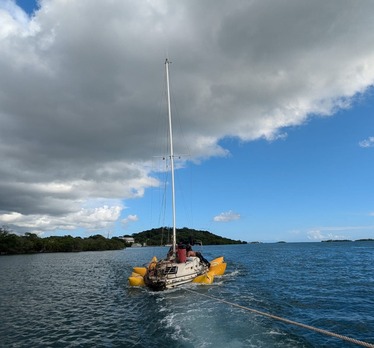  A sailboat is surrounded by large, yellow floats while it is towed by a line in the ocean on a sunny day with clouds.