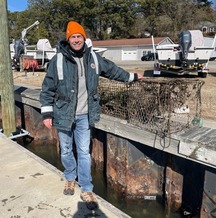 A person wearing a jacket and an orange beanie stands on a dock in the sun holding two retrieved crab trap debris items.
