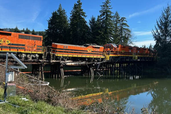 One of several orange Portland and Western train engines derailed from a damaged bridge rests between two upright engines.