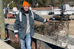 A person wearing a jacket and an orange beanie stands on a dock in the sun holding two retrieved crab trap debris items.