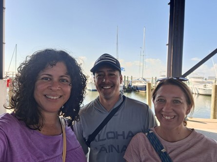 Three team members smile for a photo at a harbor with vessels and water behind them.