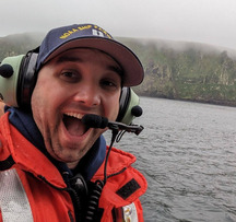 A man wearing a NOAA Ship Fairweather cap, life vest, and headset smiles on a boat in front of a misty coastal cliff.