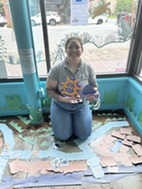 A person crouches in a homemade paper salt marsh, holding craft items