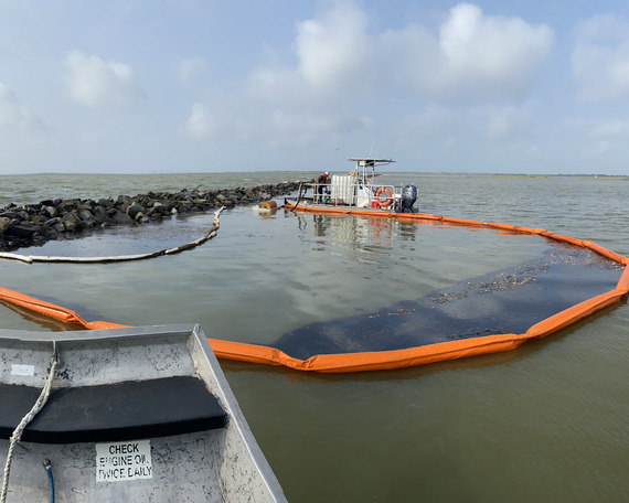 View from a response boat showing dark oil contained within an orange floating boom barrier near a rock jetty and a second work boat. 
