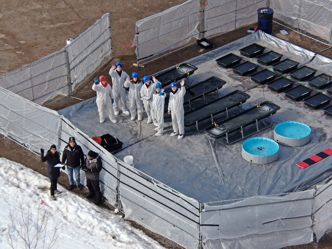 Aerial view of researchers in protective suits and hard hats waving next to testing troughs, plastic pools, and equipment.