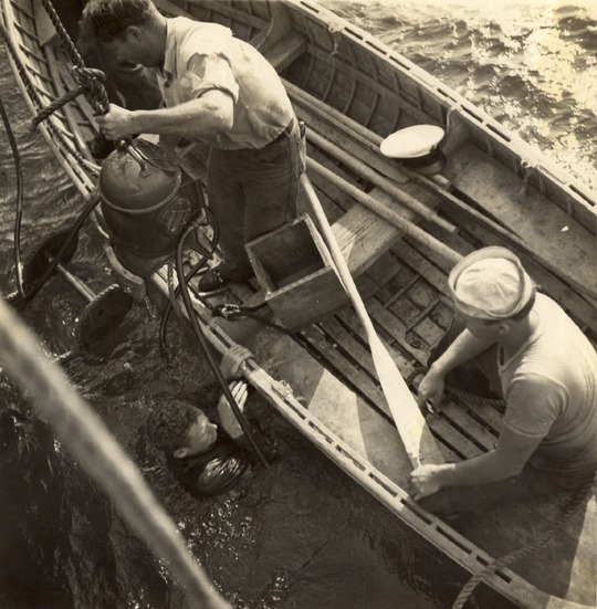 A man in a wooden boat lowers a vintage diving helmet to a diver submerged in the water