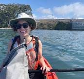 A woman wearing a sun hat and life jacket smiles while paddling an orange kayak on open water.