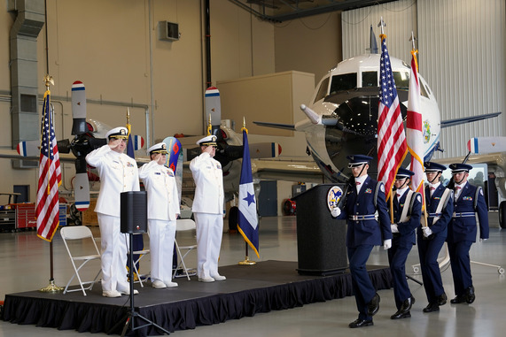 Officers on stage salute a color guard as it passes in front of them and a NOAA aircraft