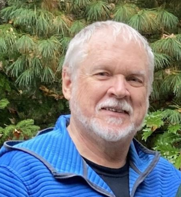 older man with white beard in front of green trees in a blue sweater 