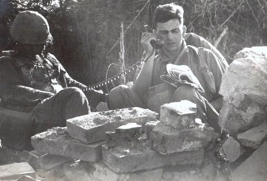 Black and white photo of two marines sitting on the ground, one looking at a manual and holding a phone receiver