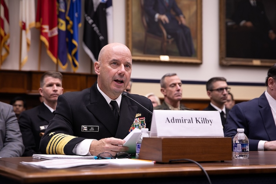 man in navy uniform seated at table, holding papers, speaking into a microphone