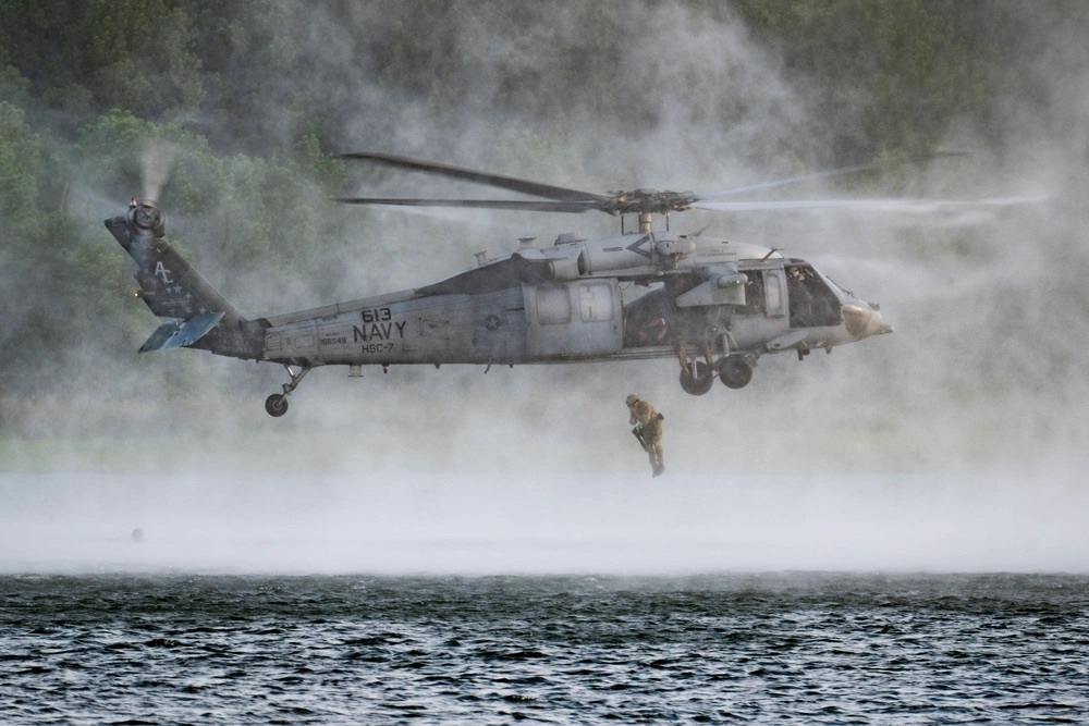 helicopter over grey water with cloudy background with person in yellow hanging underneath