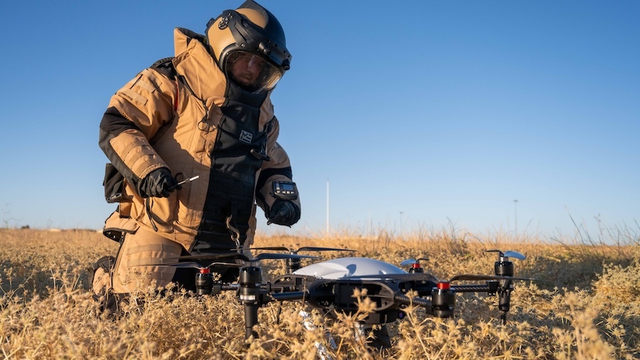man in heavy protective gear and helmet kneeling over a black quad rotor drone in a field of yellow grass