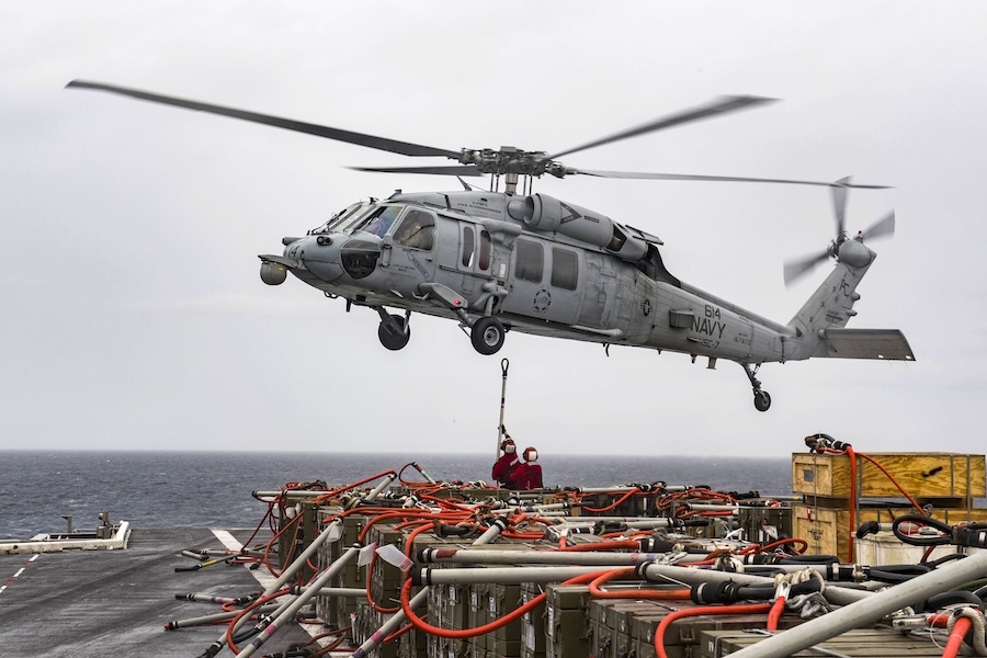 helicopter hovering over a aircraft carrier flight deck covered with creates. two people in red stand below holding a stick with loop at the end