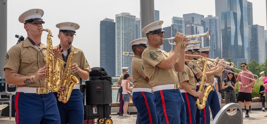 Marines in brown shirts and blue pants play brass instruments on a waterfront, tall buildings in background