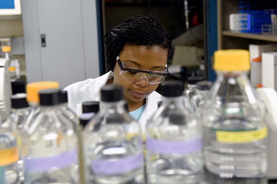 woman in safety googles looking down, row of glass bottles in front of her