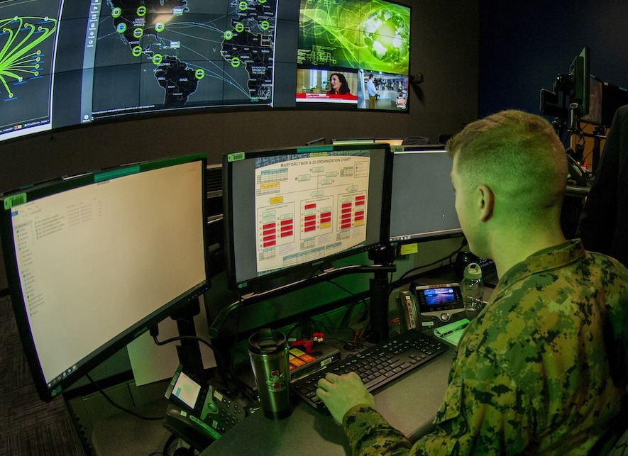 Marine in fatigues in front of a bank of computer screens glowing green