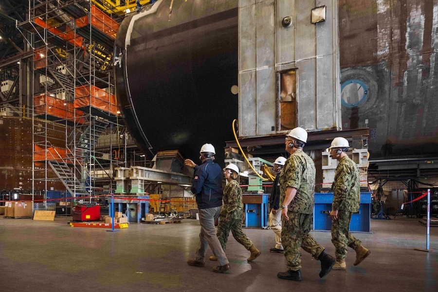 a group of people walks in front of an elevator submarine hull with tall scaffolding just beyond