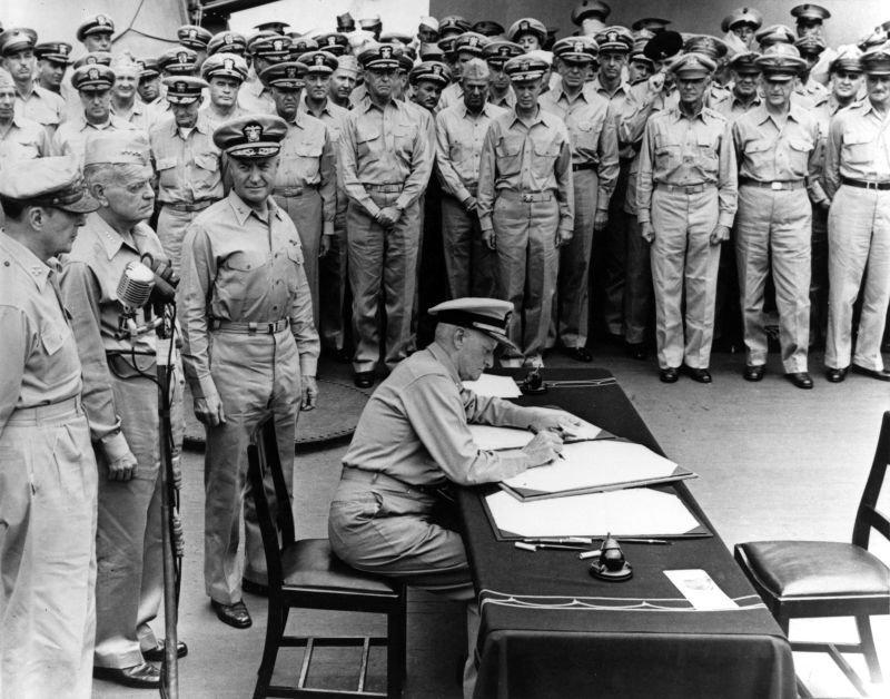 nimitz seated at a table, signing documents, officers around him