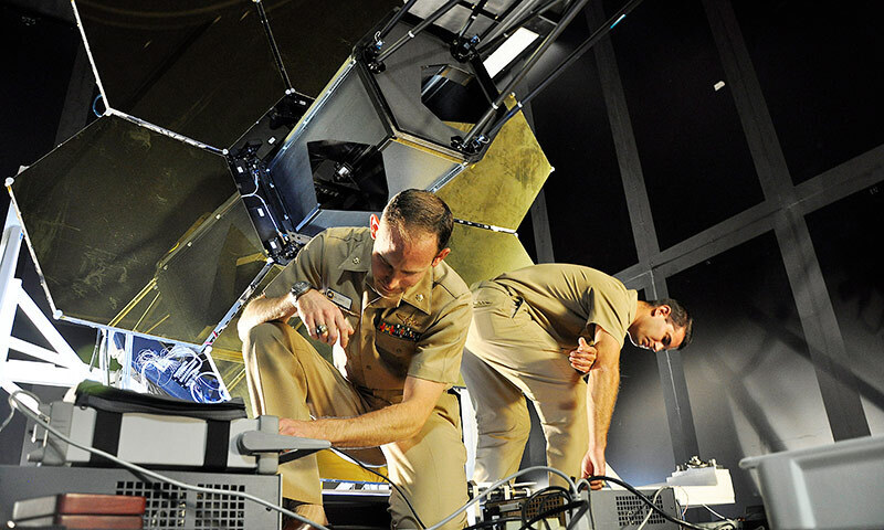 two men in uniform working on equpment benath a large satellite