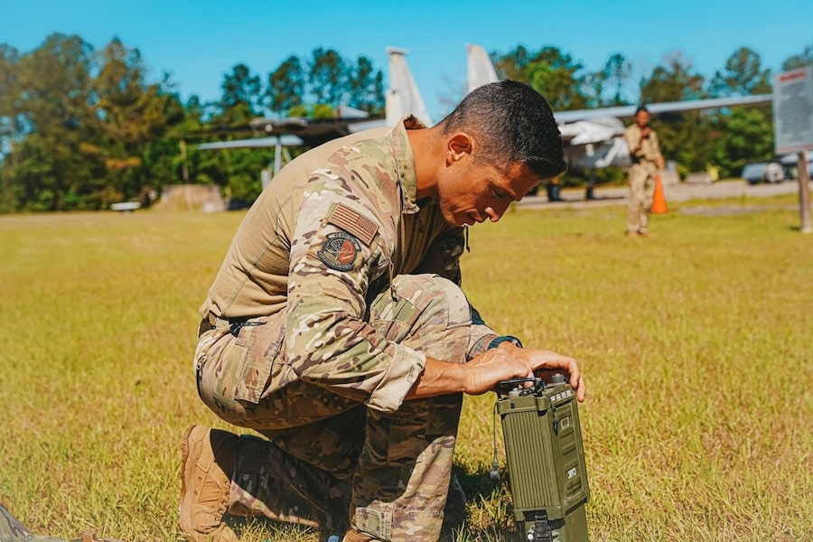 Man in camouflage kneels in a grassy field, examining a piece of electronic equipment