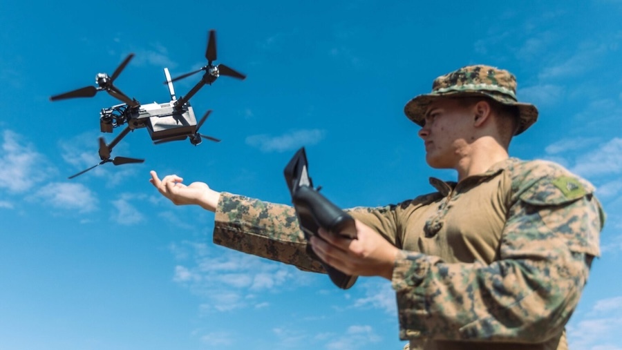 man in camouflage, hand extended, with drone hovering above