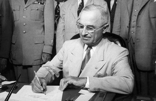 B&W photo of Harry Truman, seated at a desk, signing a document.