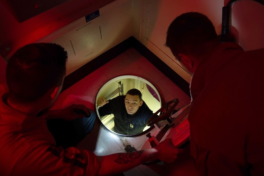 man climbing up through a hatch, two people waiting above