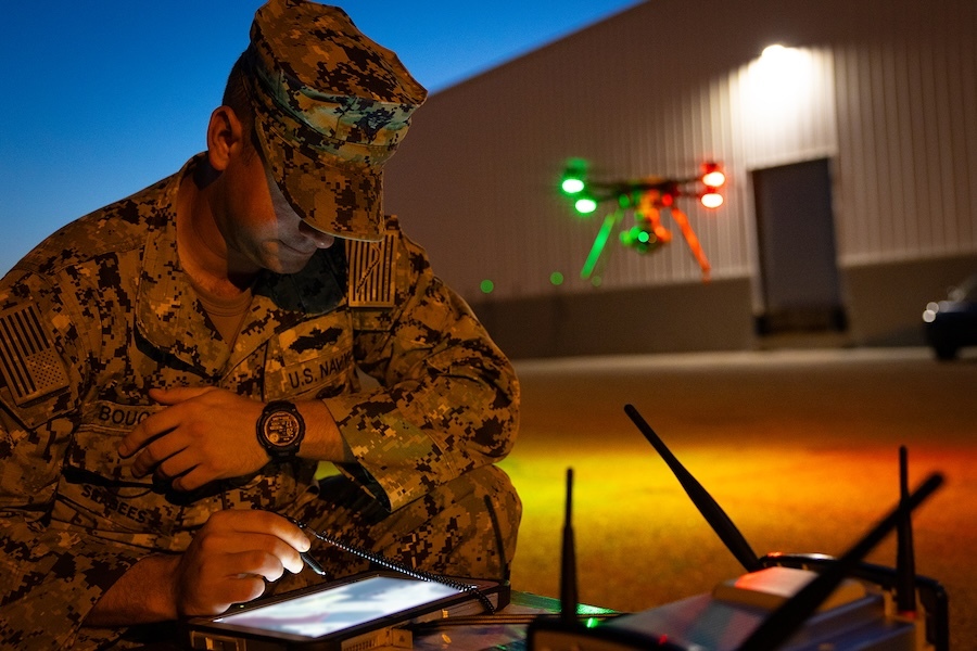 man in fatigues works on a table, drone hovers behind him