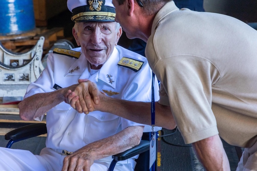 Retired Rear Adm. Bryan Compton, seated in dress whites, shakes hands with someone bending to greet him