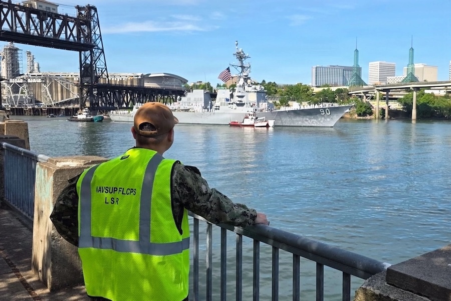 man in NAVSUP vest watches a ship enter a harbor