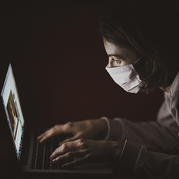 A woman in a mask watches her laptop screen intently.
