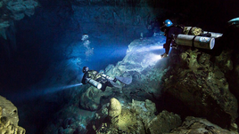 Two divers swim through a dark underwater cave in search of fossils.