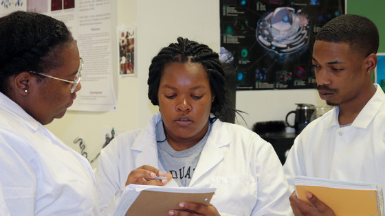 A researcher talks with two students in a laboratory settings. All are wearing lab coats.